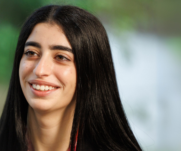 A close-up of a team member with long straight hair wearing a company lanyard, in a bright workspace with greenery visible in the background.