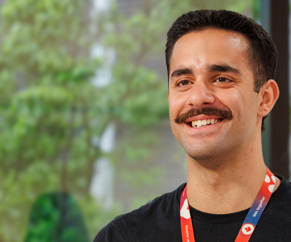A team member wearing a black shirt and company lanyard, standing in front of large windows with green foliage outside.