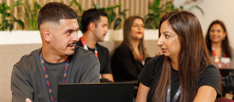 Two colleagues sitting side by side in a modern office lounge area, engaged in conversation with laptops in front of them. Other team members are visible in the background, creating a collaborative and open work environment with greenery and wooden accents.