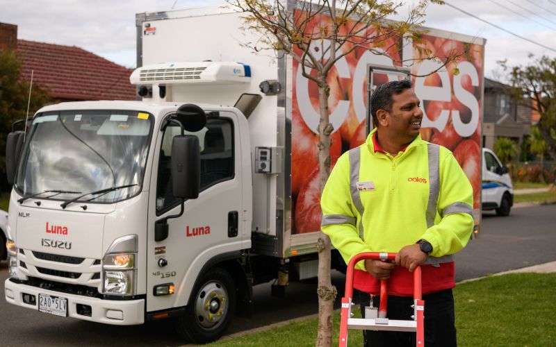 A male delivery driver wheels a trolley outside a house, with a Coles van parked on the street.