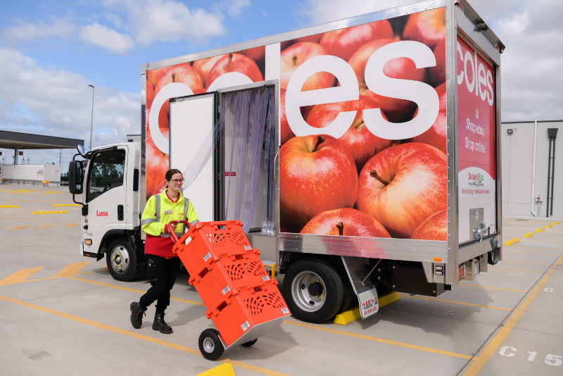 A Coles delivery truck with large apple graphics parked in a loading area. A person wearing a high-visibility jacket is moving bright orange crates on a trolley near the open rear door of the truck.