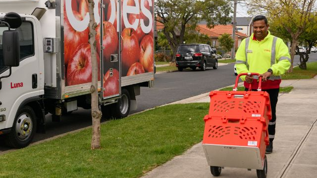 Male delivery driver wheeling in two totes on a trolley in the street with the Coles delivery van parked in the background