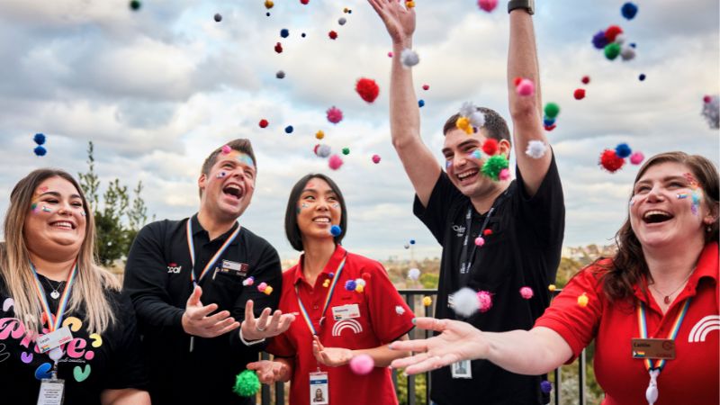 A group of Coles team members outdoors tossing colourful pom-poms into the air, wearing Coles lanyards and uniforms. The image reflects teamwork, celebration, and an inclusive, fun workplace culture.