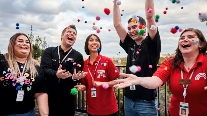A group of five team members, three females and two males, are celebrating pride with colorful balls in the air. They are all smiling and looking up at the balls with amusement