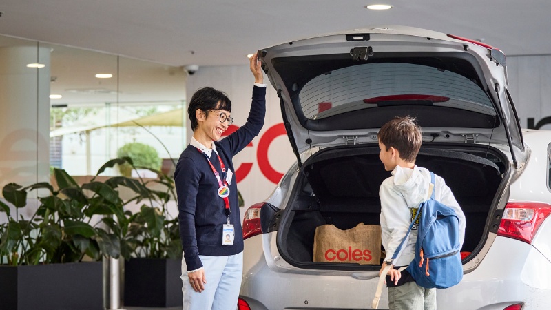 A Coles team member standing beside an open car boot with a Coles shopping bag inside, greeting a child wearing a backpack. The image represents work-life balance, flexibility, and family-friendly values supported by Coles.