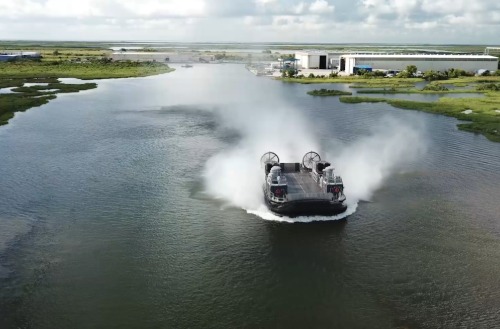Textron Systems hovercraft traveling across a coastal waterway near an industrial marine facility.