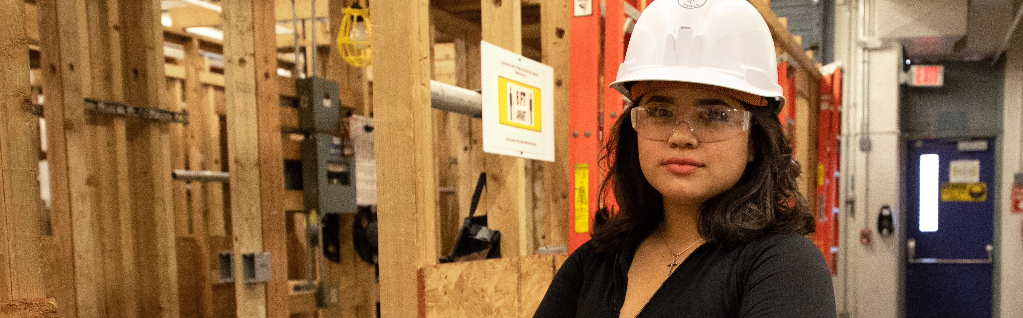 Female skilled trades student wearing protective gear standing next to a framed wall