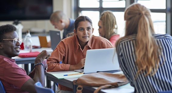 3 students sit around a table talking together. One student in the middle has their laptop open in front of them.