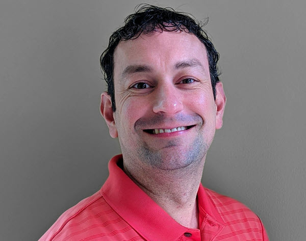 Jon R. smiling at the camera. He has short, brown hair and a bright, coral collared shirt.