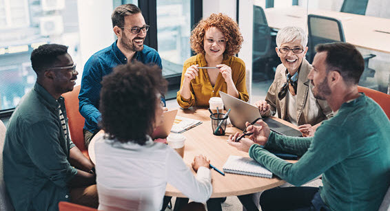 A group of employees sitting around a table in a conference room. They are smiling and all looking at one man who is talking to them.