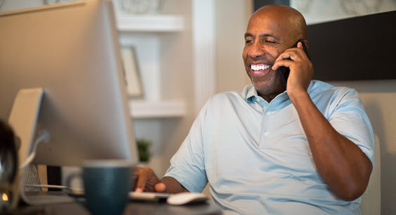 A man is sitting in front of his computer, while talking on his cell phone.