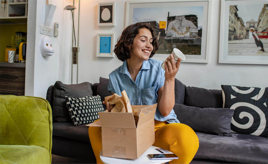 Photo of a woman sitting on a couch and admiring a mug as she unpacks it from a brown shipping box.