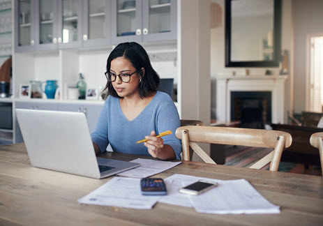 Woman at the kitchen table reviewing her finances on her laptop computer